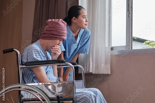 Uniformed young Asian female therapy doctor with wheelchair male patient at window to support recovery  suffer and cough from cancer illness after chemo medical treatment in hospital inpatient room.