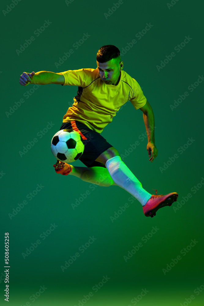 Studio shot of young professional male football soccer player in motion isolated on green background in neon. Concept of sport, goals, competition, hobby, achievements