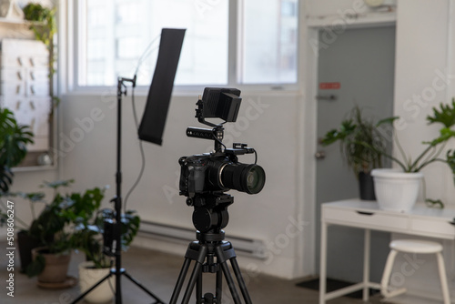 Video Camera with External Monitor and Cage on Tripod with Lighting in Background. Behind the Scenes on Commercial Film Set in White Studio.