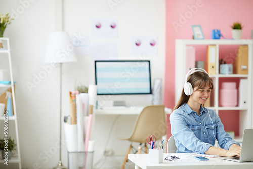 Cheerful young lady in wired headphones sitting at table and using laptop while working with pleasure on web project