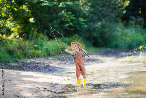 4-year-old child in wicker hat is fishing with stick, fishing rod and rope in grass by lake, boy play fishing. Happy childhood and holidays in the countryside,