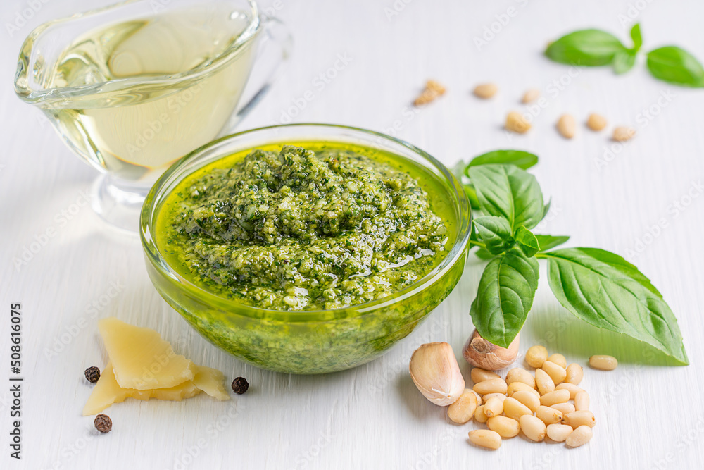 Italian homemade herbal pesto sauce made of blended parmesan cheese, green basil leaves, pine nuts, garlic, black pepper and olive oil served in glass bowl on white wooden background used on pasta Italian homemade herbal pesto sauce made of blended parmesan cheese, green basil leaves, pine nuts, garlic, black pepper and olive oil served in glass bowl on white wooden background used on pasta