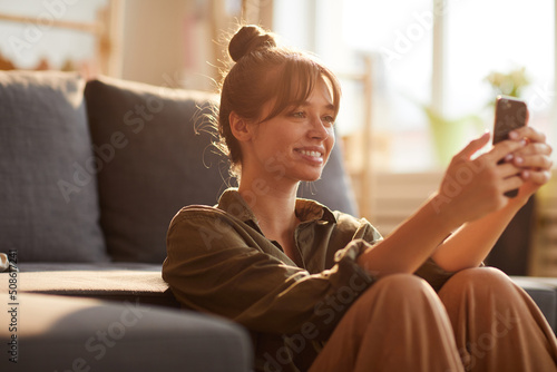 Smiling beautiful girl with bangs leaning on sofa and browsing Internet on smartphone while searching for information