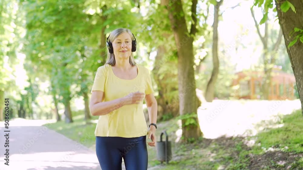 Senior active gray-haired woman jogging in urban city summer park outdoor. Mature old fitness female running exercise with headphones enjoys and listens to music. Elderly sport runner Workout cardio Senior active gray-haired woman jogging in urban city summer park outdoor. Mature old fitness female running exercise with headphones enjoys and listens to music. Elderly sport runner Workout cardio