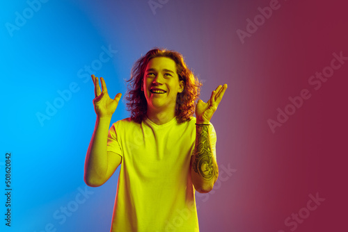 Young attractive man with long curly hair posing isolated on gradient blue-pink background. Concept of beauty, fashion, youth culture and emotions