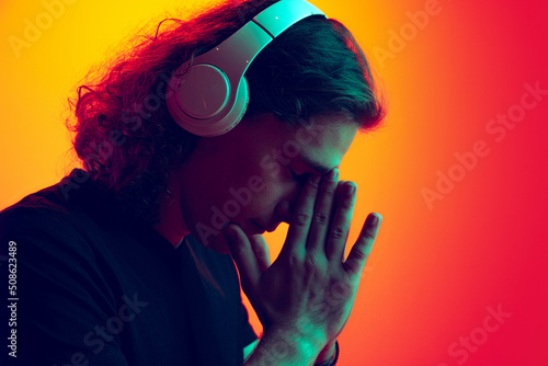 Young excited man with long curly hair listening to music in headhones isolated on gradient yellow-pink background in neon. Concept of beauty, fashion, youth culture and emotions © master1305