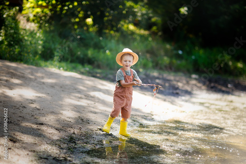 Preschooler boy in yellow rubber boots plays fishing, child fishes with stick near lake in countryside, games in nature and childhood without gadgets, summer holidays © natalialeb