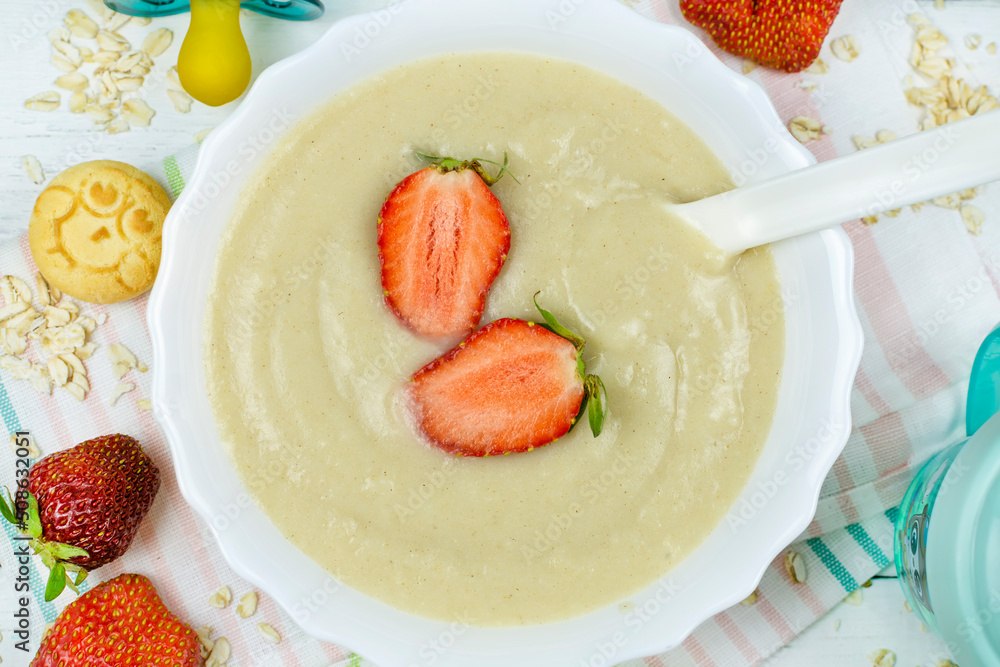 Oatmeal porridge for a baby with strawberries in a white bowl with a spoon, cookies, a drinking cup, a dummy close-up. The first complementary food of a child, baby nutrition. Oatmeal porridge for a baby with strawberries in a white bowl with a spoon, cookies, a drinking cup, a dummy close-up. The first complementary food of a child, baby nutrition.