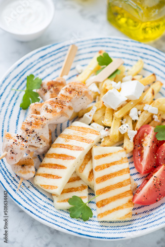 Slices of grilled haloumi cheese, souvlaki skewers, greek-style fries and tomatoes served on a blue and white plate, vertical shot, close-up