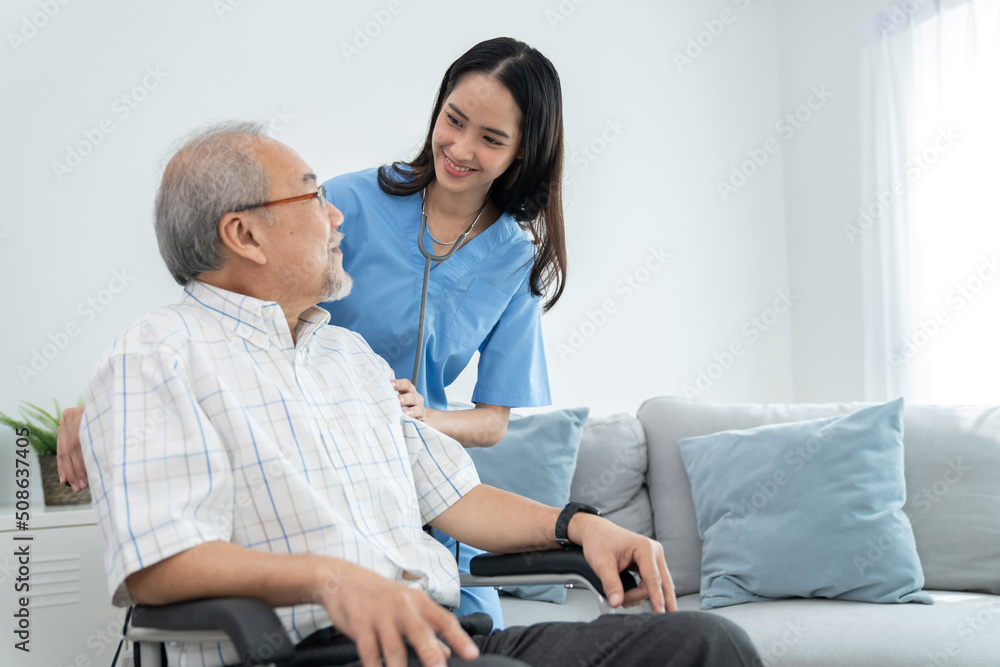 Obraz premium Attractive caregiver nurse stand behind elderly man who sitting on the wheelchair and smile at home. Doctor take care and encourage patient. Rehabilitation and healthcare concept