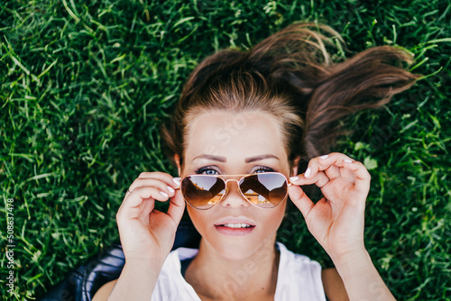 Close up portrait of beautiful young woman laying on green grass and looking over her aviator sunglasses holded by her hands. Summer time concept. © Iona