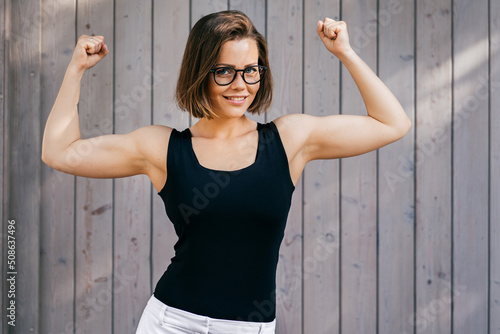 Strong fitness girl with short hair dressed casual, demonstrates her biceps outdoor over wooden background