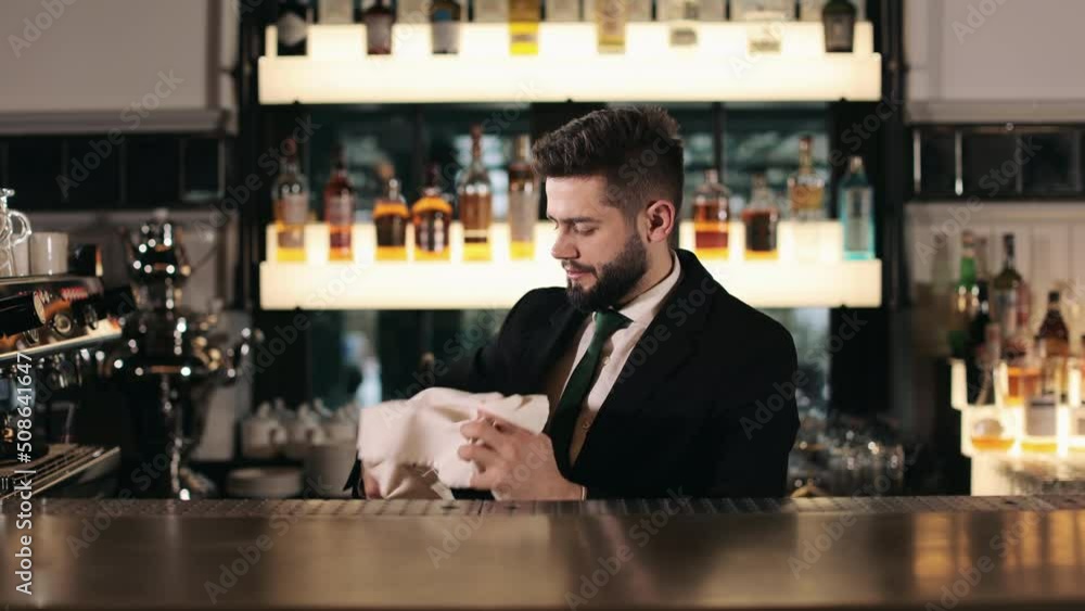 Handsome young bartender wiping wine glass with towel behind bar counter. Caucasian man in black suit preparing silverware for serving at modern restaurant. Handsome young bartender wiping wine glass with towel behind bar counter. Caucasian man in black suit preparing silverware for serving at modern restaurant.