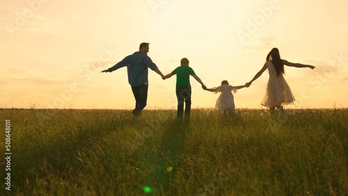 Big happy family is walking on summer field holding hands. Joyful parents, children playing running in park at sunset. Family walks on green grass in meadow. Family walk in nature. Young family in sun