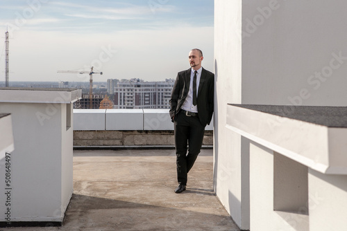 a man in a black suit with a tie leaned against the wall on the roof against the background of the panorama of the city and building cranes