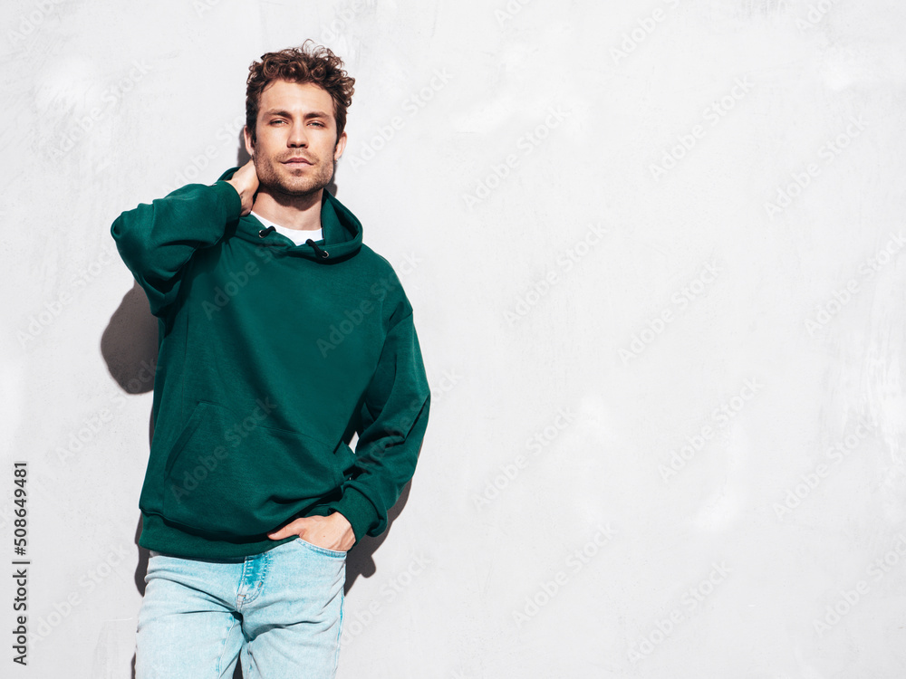 Handsome confident hipster model.Sexy unshaven man dressed in summer stylish green hoodie and jeans clothes. Fashion male with curly hairstyle posing in studio. Isolated on grey Handsome confident hipster model.Sexy unshaven man dressed in summer stylish green hoodie and jeans clothes. Fashion male with curly hairstyle posing in studio. Isolated on grey