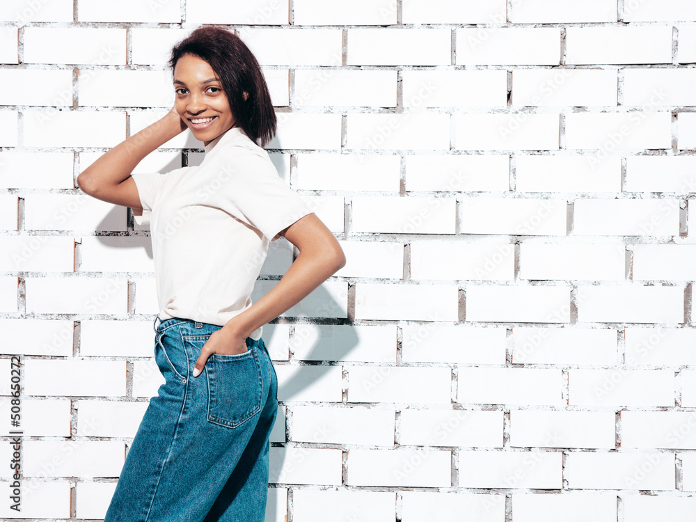 Portrait of young beautiful black woman. Smiling model dressed in summer jeans clothes. Sexy carefree female posing near white brick wall in studio. Tanned and cheerful Portrait of young beautiful black woman. Smiling model dressed in summer jeans clothes. Sexy carefree female posing near white brick wall in studio. Tanned and cheerful