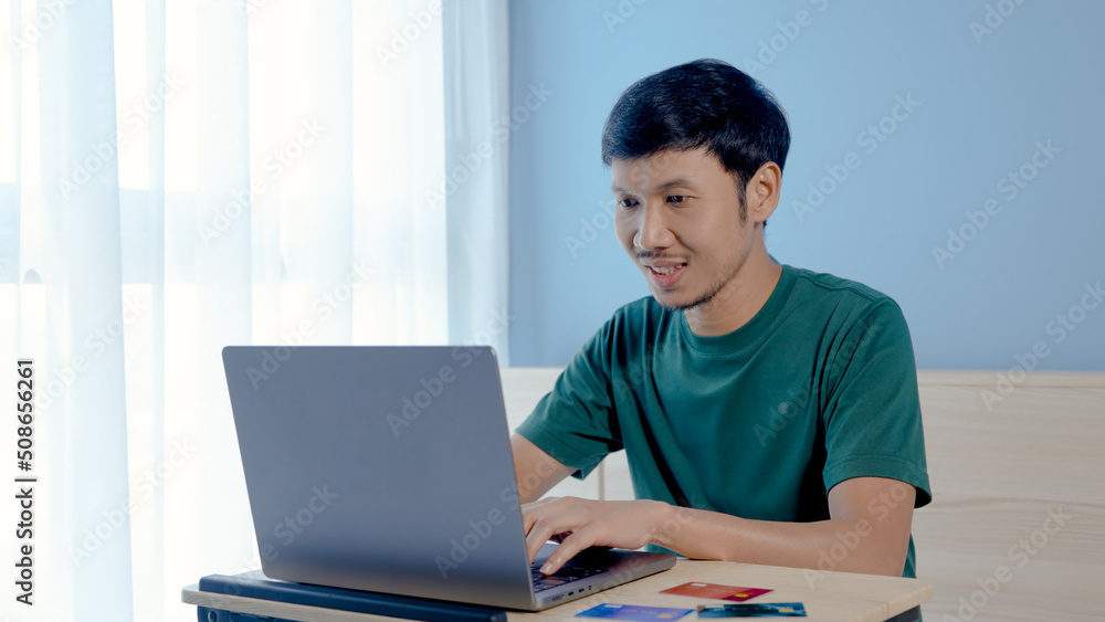 Handsome Asian man wearing a yellow shirt pays for online shopping on his laptop computer. The young man looked happy after completing his credit card payment.online shopping Handsome Asian man wearing a yellow shirt pays for online shopping on his laptop computer. The young man looked happy after completing his credit card payment.online shopping