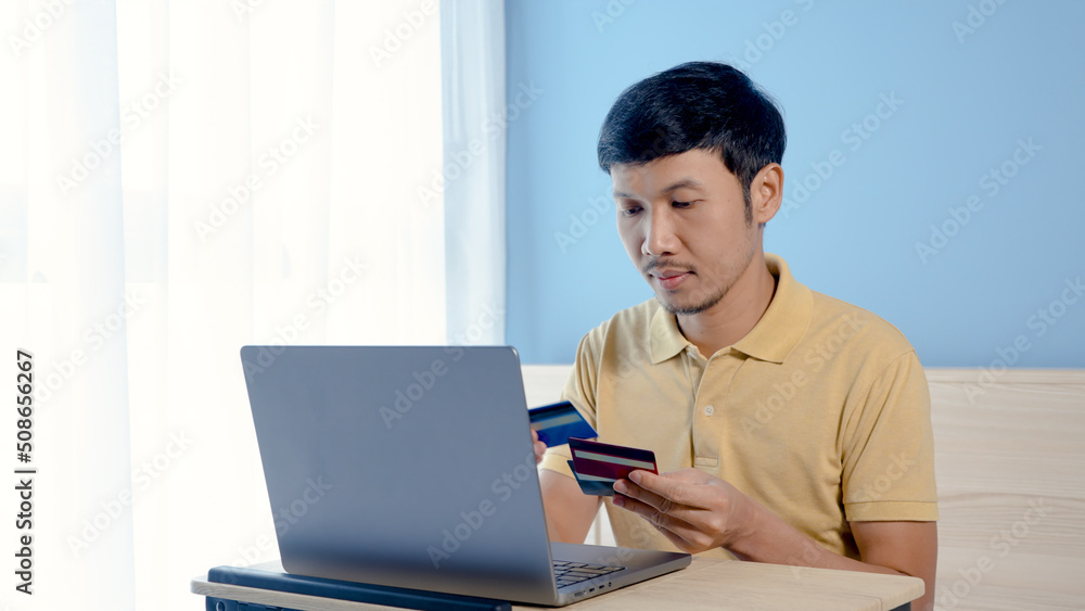 Handsome Asian man, wearing a yellow shirt sitting on a white bed, has a serious look on his face when he chooses a credit card to pay for an order online, on his laptop computer.. Handsome Asian man, wearing a yellow shirt sitting on a white bed, has a serious look on his face when he chooses a credit card to pay for an order online, on his laptop computer..