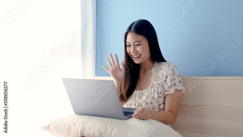 Beautiful Asian girl, wearing a white floral shirt, is video call chatting with friends on a laptop computer, on vacation chilling in his bedroom, greeting and chatting happily.