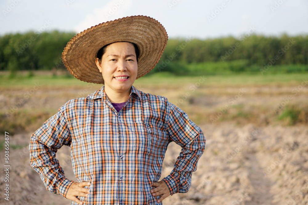 Portrait of Asian middle-aged woman farmer is at agriculture plantation, wears hat, plaid shirt, smile and put hands on hips, feels confident. Concept  : happy farmer. Agriculture occupation. 