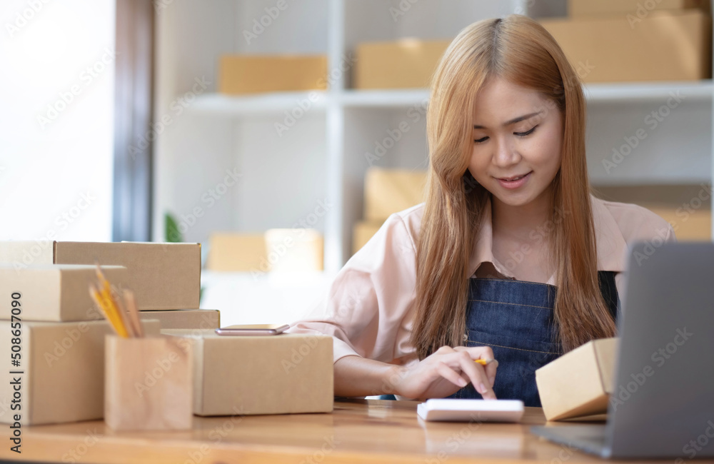 Startup small business SME. Beautiful asian entrepreneur using calculator, check online order and prepare to packing for customer. Successful SME business online concept. Startup small business SME. Beautiful asian entrepreneur using calculator, check online order and prepare to packing for customer. Successful SME business online concept.