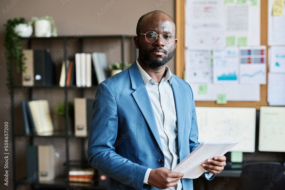 Portrait of African confident businessman in eyeglasses and blue suit looking at camera while working with documents at office Portrait of African confident businessman in eyeglasses and blue suit looking at camera while working with documents at office