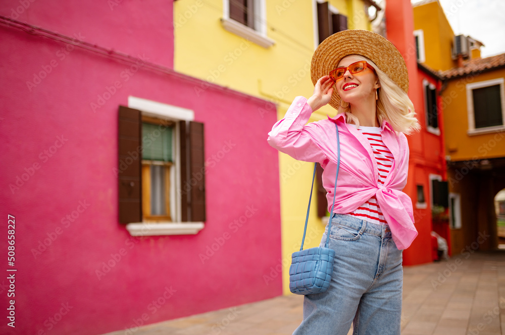 Happy smiling traveler woman walking in street with pink, yellow houses. Model wearing trendy summer outfit with orange sunglasses, straw hat, shirt,  shoulder blue bag. Copy, empty space for text