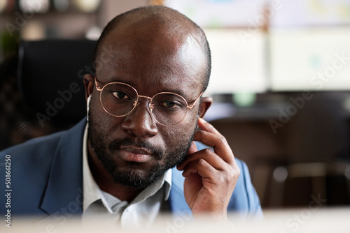 Close-up of African serious businessman in eyeglasses using wireless headphones for conversation online