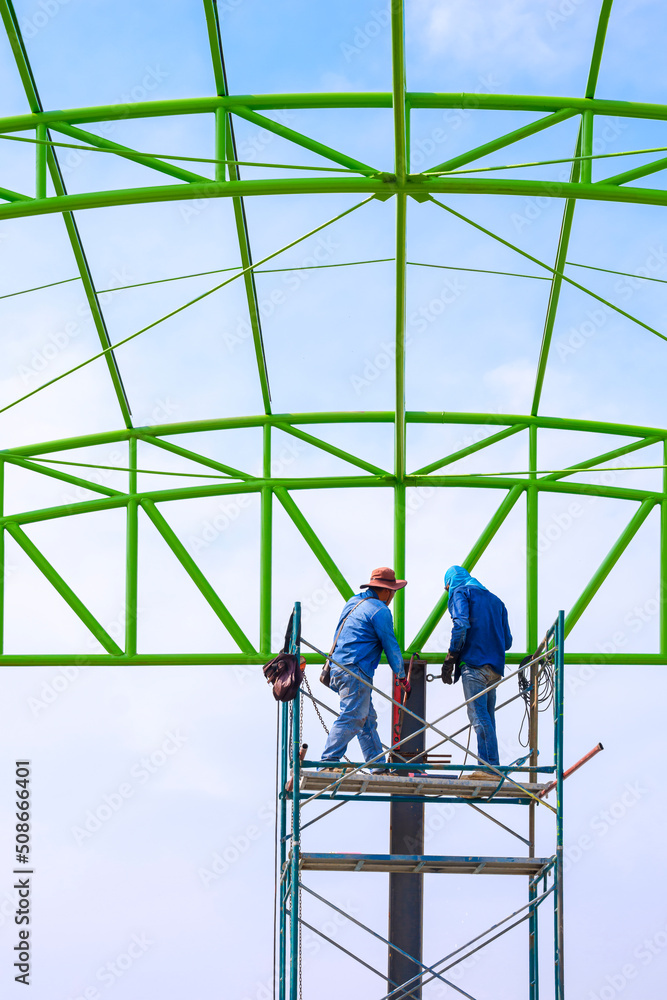 Asian foreman and construction worker on scaffolding are working to build metal roof structure of warehouse building in construction site