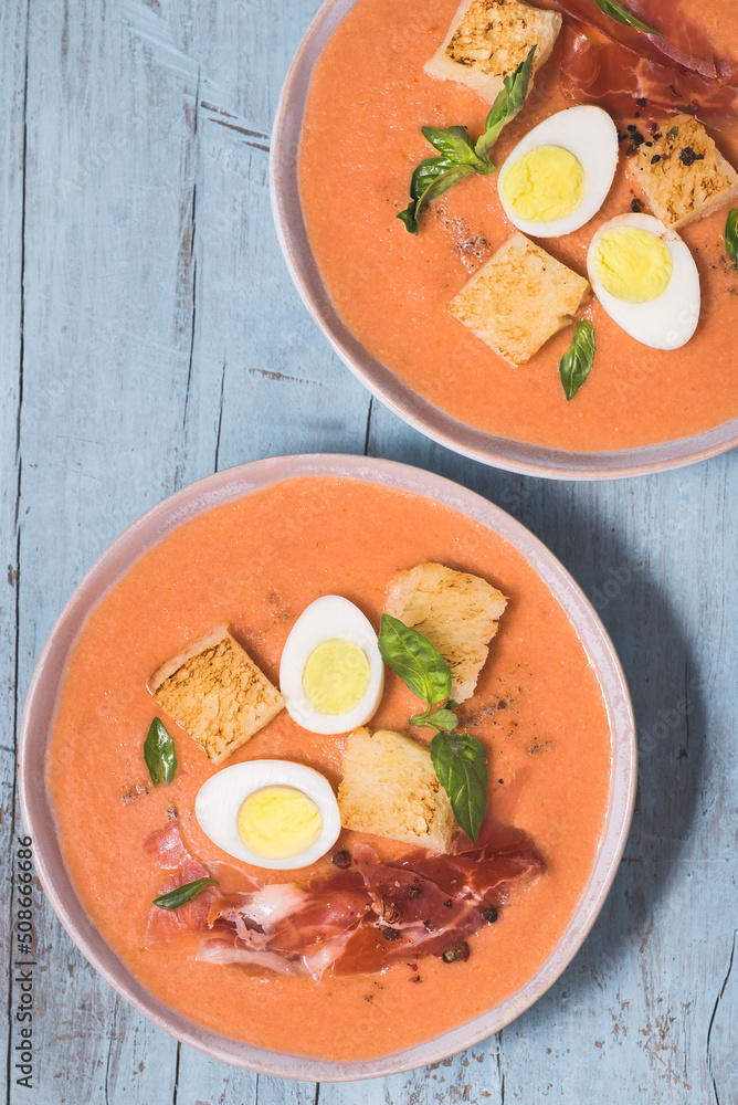 Spanish salmorejo soup, tradition Andalusian
cold tomato soup topped with serrano ham (jamon), boiled egg, bread and  pepper on the blue wooden background. Summer recipes, menu, close-up