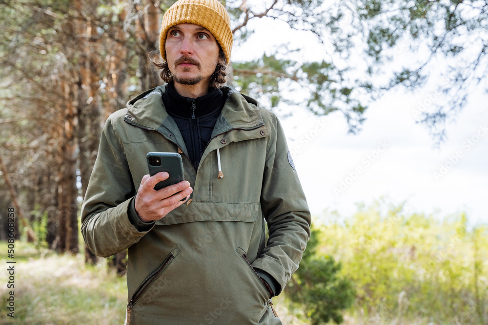 A bearded guy holds a smartphone in his hands, a man with a phone in his hands, a man standing in the woods, a pensive look, a hipster on a hike in the mountains.