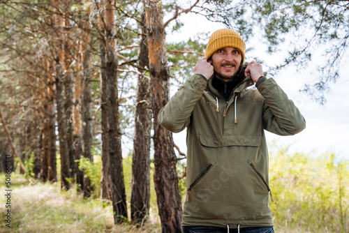 The guy is smiling standing in the forest, a hat on his head, a warm windbreaker from the rain, a man puts a hood on his head, a tourist on a hike in a pine forest.