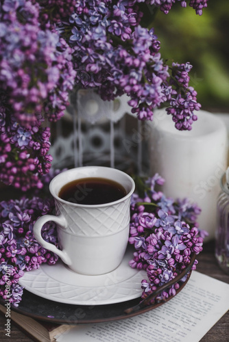 A beautiful postcard. A white coffee cup with a saucer, candles and a vase with a bouquet of purple lilac. The color 2022 is very peri. Beautiful still life. Spring time. The concept of "Good morning"