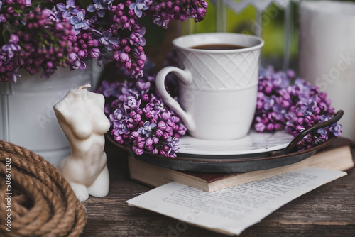 A beautiful postcard. A white coffee cup with a saucer, candles and a vase with a bouquet of purple lilac. The color 2022 is very peri. Beautiful still life. Spring time. The concept of "Good morning"
