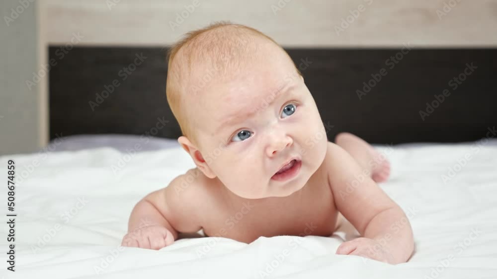 Cute baby girl frowns trying to raise up head lying on bed. Curious newborn kid examines bedroom looking around and opening mouth close view Cute baby girl frowns trying to raise up head lying on bed. Curious newborn kid examines bedroom looking around and opening mouth close view