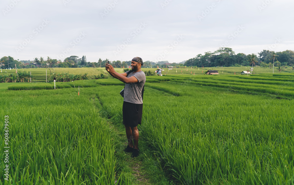 Emotional hipster guy in casual bandanna using cellular device for clicking scenery view during summer trip to Asia, happy male blogger with smartphone gadget shooting video content at rice fields