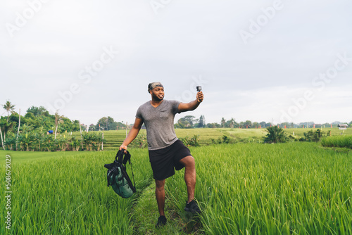 African American man using go pro camera for shooting influence vlog during travel journey in Indonesia for walking around rice fields, hipster guy with backpack clicking images via waterproof device photo