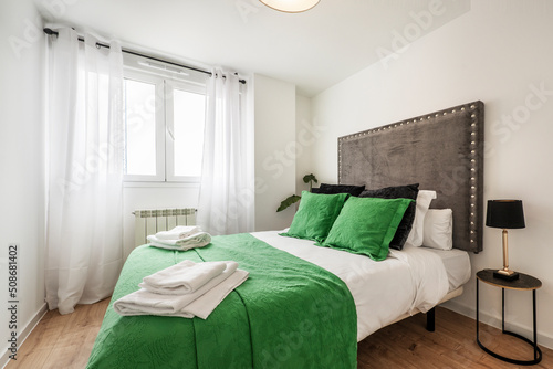 Bedroom with a double bed dressed in a striking green bedspread and matching cushions, upholstered fabric headboard and white curtains on the window