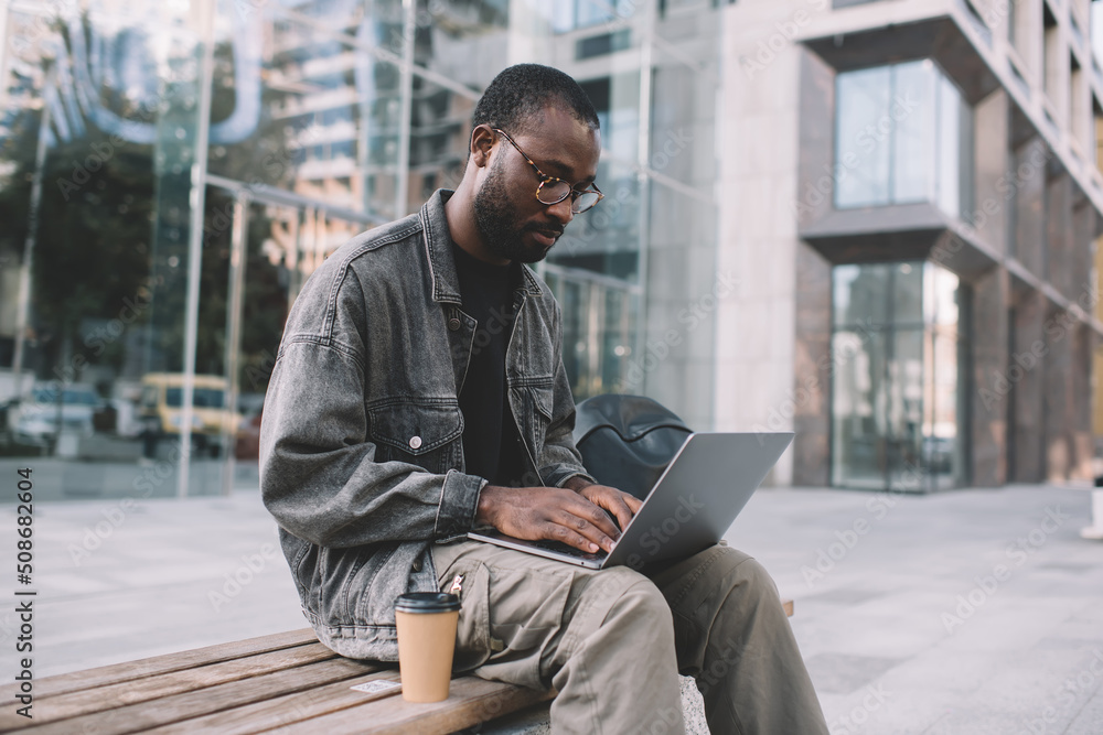 Skilled male programmer in classic eyewear browsing website for editing graphic design via media application on modern laptop, millennial African American software developer working remotely in city Skilled male programmer in classic eyewear browsing website for editing graphic design via media application on modern laptop, millennial African American software developer working remotely in city
