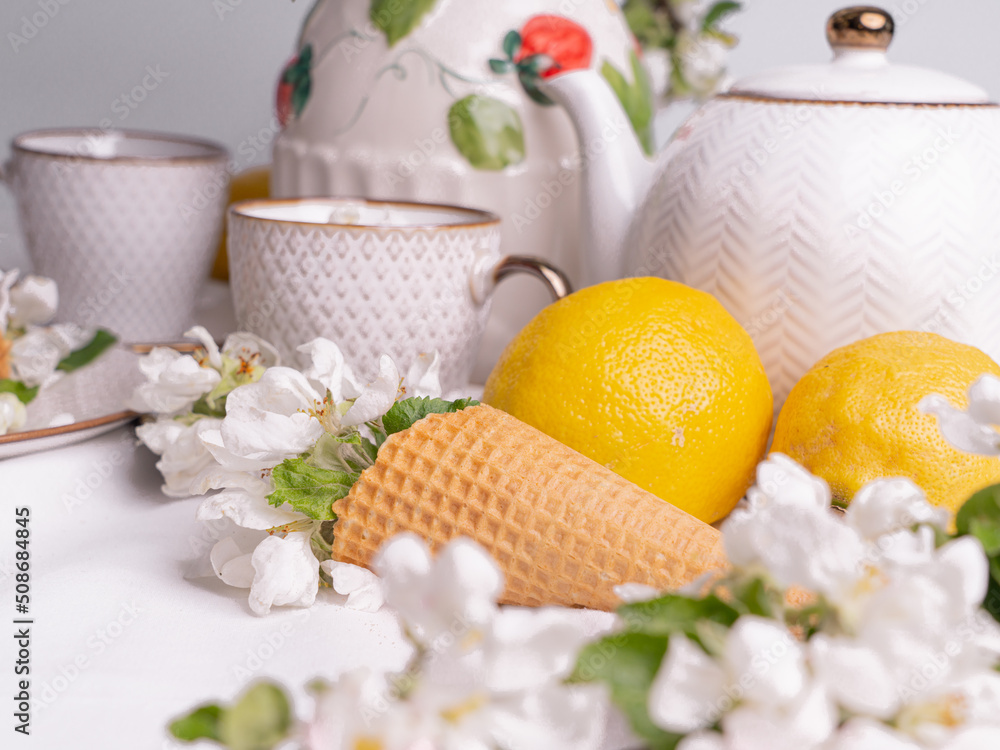 White tea set with tea in cups with yellow lemons and a blossoming apple branch.Close-upThere are waffle cones on a saucer.