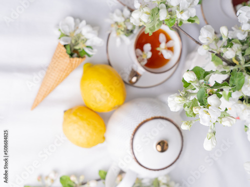 White tea set with tea in cups with yellow lemons and a blossoming apple branch.View from aboveThere are waffle cones on a saucer.