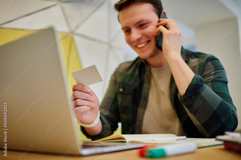 Young smiling man calling via mobile phone while sitting at the desk with laptop computer. Working online on freelance, using technology . Selective focus