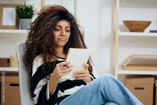 Pensive concentrated pretty beautiful curly tanned Latin woman in striped shirt hold notebook look down read work letter essay in home interior. Copy space Banner. Writer concept