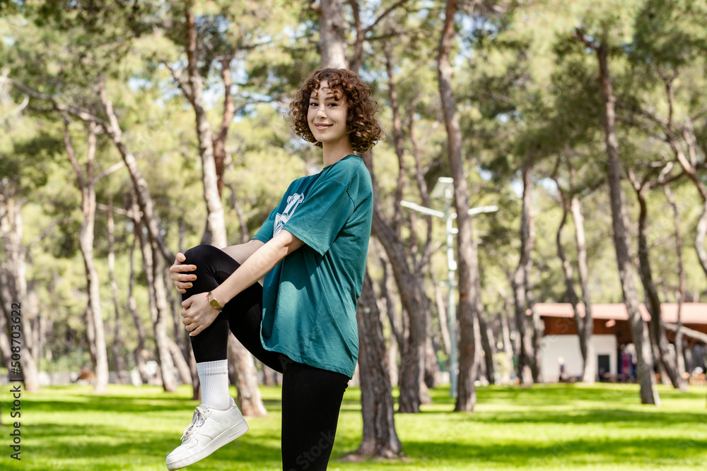 Happy young redhead woman wearing green t-shirt and black legging standing on city park, outdoors runner athlete woman doing fitness stretch. Holding one leg for stretching hamstring muscles. Happy young redhead woman wearing green t-shirt and black legging standing on city park, outdoors runner athlete woman doing fitness stretch. Holding one leg for stretching hamstring muscles.