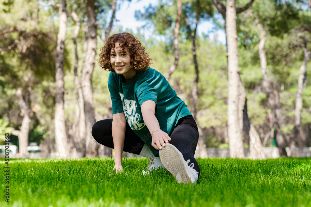 Beautiful young redhead woman wearing sportive clothes standing on city park, outdoors stretching leg muscles, doing side lunges in skandasana pose. Healthy lifestyle concept. Beautiful young redhead woman wearing sportive clothes standing on city park, outdoors stretching leg muscles, doing side lunges in skandasana pose. Healthy lifestyle concept.
