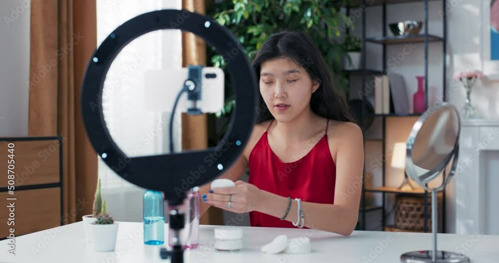 Removal makeup. Woman cleansing her face with a white cotton pad, looking at herself in the mirror. Influencer recording beauty tutorial skin care vlog using ring light phone for social media channel. Removal makeup. Woman cleansing her face with a white cotton pad, looking at herself in the mirror. Influencer recording beauty tutorial skin care vlog using ring light phone for social media channel.