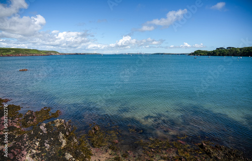 looking across a beach with shallow water and seaweed foreground across a large turquoise blue estuary under a blue sky with white cloud