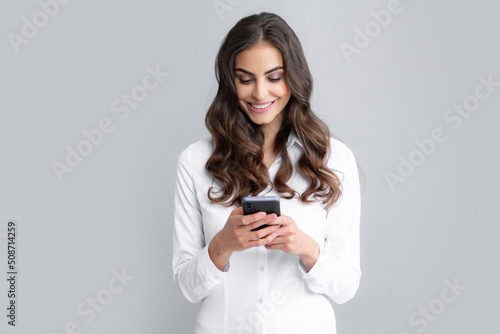 Young woman reading message on mobile phone and smiling. Happy businesswoman with smartphone, standing on grey background.