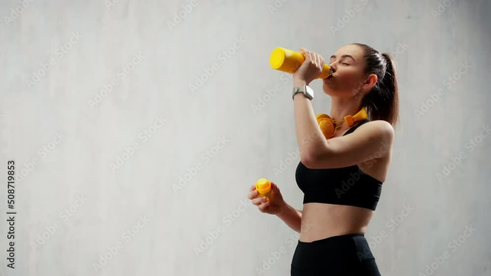 Young sporty woman in sportswear with wireless headset drinking from bottle over grey background Young sporty woman in sportswear with wireless headset drinking from bottle over grey background